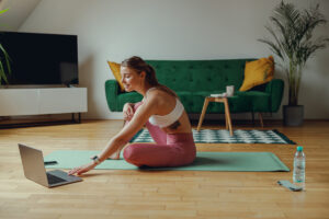 Woman on yoga mat in sportswear, using laptop on hardwood flooring